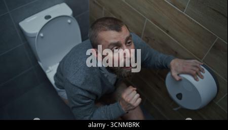 Homme d'affaires avec expression faciale tendue souffrant de diarrhée et d'indigestion, assis sur les toilettes. Homme éprouvant des maux d'estomac, inconfort digestif dans les toilettes du bureau pendant le travail. Angle élevé Banque D'Images