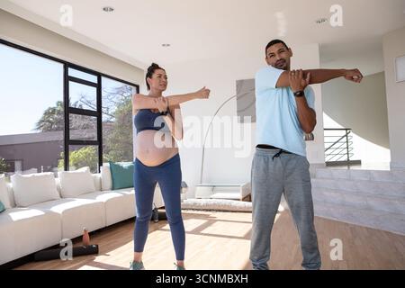 Couple diversifié étirant les bras sur des tapis de yoga dans le salon avec une femme enceinte tenant une bouteille d'eau Banque D'Images