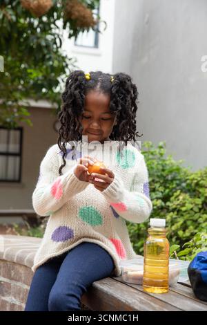 Enfant afro-américaine pelant l'orange sur le rebord dans la cour avec bouteille de jus, sandwich Banque D'Images