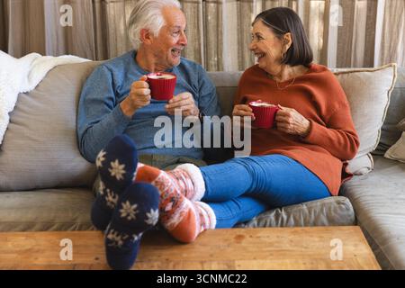 Couple senior assis sur un canapé rembourré beige dans un salon confortable tenant des tasses côtelées rouges Banque D'Images