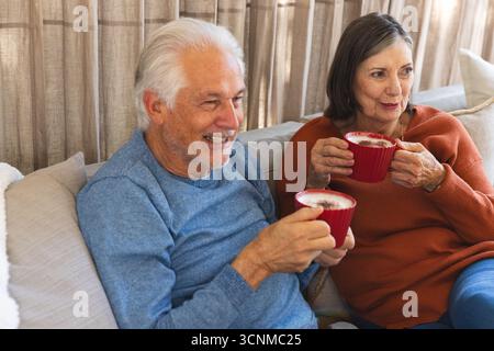 Couple senior assis sur un canapé beige tenant des tasses en céramique rouge dans le salon avec des oreillers décoratifs Banque D'Images