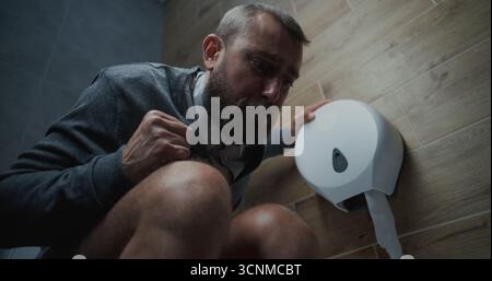 Homme avec expression faciale tendue assis sur les toilettes, souffrant de diarrhée et estomac Ache dans les toilettes. Homme d'affaires éprouvant l'inconfort et l'indigestion pendant le travail dans le bureau moderne. Angle bas. Banque D'Images