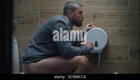 Homme assis sur les toilettes et souffrant de diarrhée et estomac Ache dans les toilettes. Homme d'affaires éprouvant l'inconfort, l'indigestion et l'inconfort digestif pendant le travail dans le bureau moderne. Prise de vue moyenne. Banque D'Images