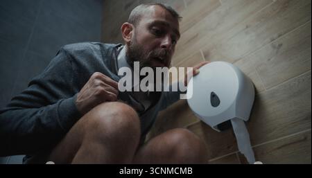 Homme avec expression faciale tendue assis sur les toilettes, souffrant de diarrhée et estomac Ache dans les toilettes. Homme d'affaires éprouvant l'inconfort et l'indigestion pendant le travail dans le bureau moderne. Angle bas. Banque D'Images
