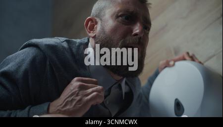 Homme d'affaires avec expression faciale tendue souffrant de diarrhée et d'indigestion, assis sur les toilettes. Homme éprouvant des maux d'estomac, inconfort digestif dans les toilettes du bureau pendant le travail. Gros plan. Banque D'Images
