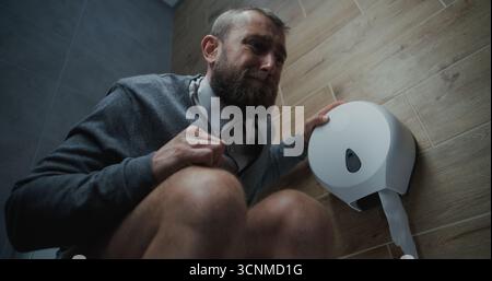 Homme avec expression faciale tendue assis sur les toilettes, souffrant de diarrhée et estomac Ache dans les toilettes. Homme d'affaires éprouvant l'inconfort et l'indigestion pendant le travail dans le bureau moderne. Angle bas. Banque D'Images