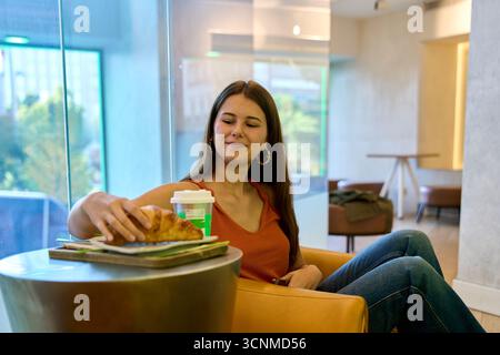 Jeune femme assise dans un café moderne, ramassant un croissant dans un plateau, avec une tasse de café à emporter à côté Banque D'Images
