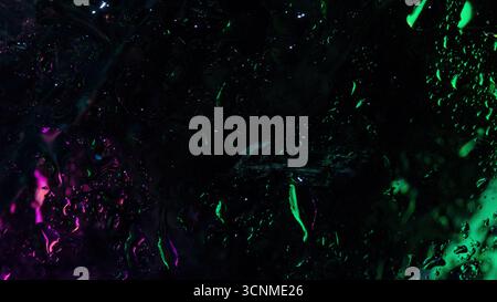 Image abstraite sombre de verre humide avec des reflets de néon vert et violet brillants, des gouttelettes d'eau et des textures tachées. Banque D'Images
