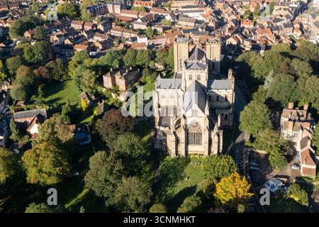 Photographie aérienne de la cathédrale de Ripon à Ripon, Yorkshire du Nord, Royaume-Uni, capturée par temps clair de fin d'été avec un ciel bleu vif et un soleil chaud mettant en valeur l'architecture gothique saisissante de la cathédrale. L'image montre l'élégant front ouest anglais ancien avec ses tours jumelles, ses pierres complexes et ses fenêtres cintrées, entouré par les rues historiques de la ville, les bâtiments voisins, les toits et les parcelles d'espaces verts, y compris les parcs et les espaces ouverts. Cette vue aérienne d'un drone capture la position centrale de la cathédrale dans la ville médiévale compacte, soulignant son rôle en tant que monument historique clé Banque D'Images