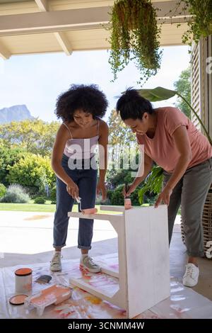 Mère diversifiée avec fille adulte peignant la table en bois avec de la peinture pêche sur le tissu de goutte sous le patio Banque D'Images