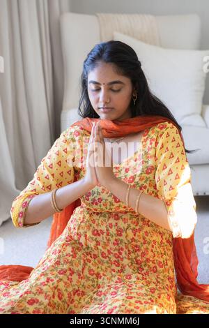 Femme indienne en kurta jaune priant avec les paumes pressées à la poitrine sur le tapis dans la chambre à coucher à la maison Banque D'Images