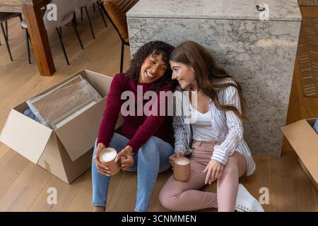 Diverses amies féminines sirotant du café tout en déballant des boîtes sur le sol par îlot de marbre dans la cuisine Banque D'Images