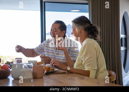 Couple senior diversifié partageant le petit déjeuner à la table à manger en bois avec panier de fruits et tasses Banque D'Images