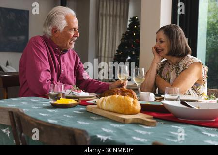 Couple senior toast repas de Noël avec verres à vin à la table à manger à la maison avec arbre de Noël Banque D'Images