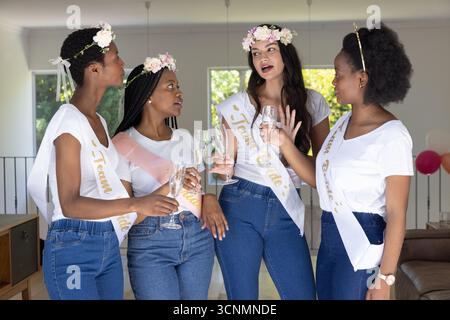 Diverses amies dégustant des flûtes à champagne portant des écharpes Team bride dans un salon moderne Banque D'Images