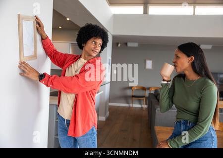 Couple diversifié accrochant cadre photo en bois et sirotant de tasse dans le salon ouvert, espace de copie Banque D'Images