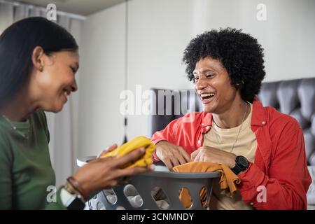 Couple diversifié rire et plier le linge à côté du lit dans la chambre avec panier, espace copie Banque D'Images