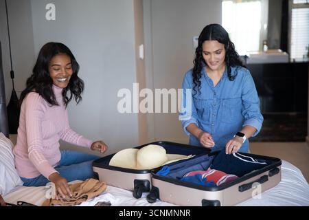 Divers amis féminins emballant la valise sur le lit à la maison avec chapeau de paille, vêtements pliés Banque D'Images
