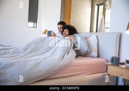 Divers partenaires masculins embrassant sous couette grise sur le lit dans la chambre tout en faisant défiler le smartphone Banque D'Images