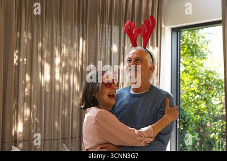 Couple senior riant et serrant dans les bras dans le salon portant des bandeaux en bois rouge et des lunettes de renne Banque D'Images