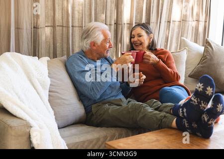 Couple senior assis sur le canapé tenant des tasses rouges à côté de la table basse en bois dans le salon Banque D'Images