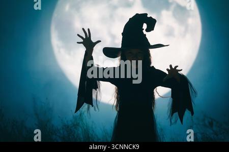 Fille vêtue de costumes de sorcière pour Halloween. Enfant debout sur fond de pleine lune. Banque D'Images