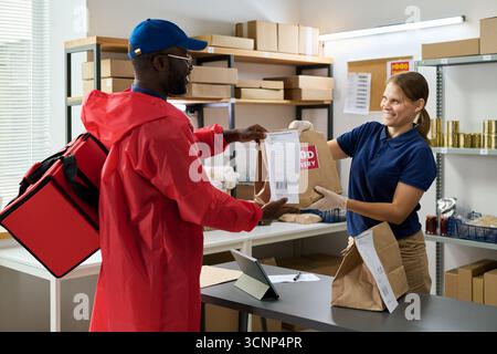 Homme noir portant un uniforme de livraison recevant une commande d'épicerie de la jeune femme adulte caucasienne souriante travaillant au comptoir dans le magasin, à la fois en interaction avec le sac en papier et le reçu imprimé Banque D'Images