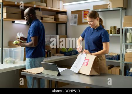 Jeune femme caucasienne adulte emballant des produits alimentaires dans un sac en papier tandis que jeune homme noir adulte triant l'épicerie dans la salle de stockage, les deux travaillant ensemble dans le centre de distribution alimentaire Banque D'Images