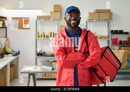 Portrait de jeune adulte Noir homme souriant et croisant les bras tout en se tenant debout dans le bureau de service de livraison, tenant un sac de livraison de nourriture isolé, portant un chapeau et des lunettes, étagères avec des paquets en arrière-plan Banque D'Images