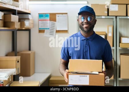 Portrait de jeune homme noir adulte portant des lunettes et un chapeau souriant tout en tenant des boîtes en carton dans la salle de stockage de l'entrepôt, debout devant les étagères avec des paquets et des colis Banque D'Images