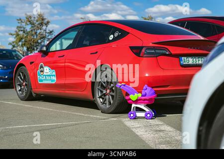 BURGAS, Bulgarie - 20 septembre 2025 : Tesla Model 3 garé à côté d'une poussette colorée sur l'asphalte sous le ciel bleu. Concept d'éco mobilité, famille Banque D'Images