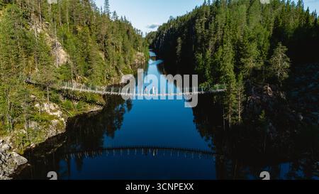Explorez la beauté naturelle époustouflante du canyon Julma Olkky dans le parc national de Hossa en Finlande. Les visiteurs traversent un pont suspendu, entouré de forêts luxuriantes et d'eaux réfléchissantes par une journée ensoleillée. Banque D'Images