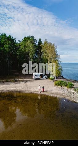 Deux personnes marchent le long de la rive calme du lac Saimaa, entouré d'arbres luxuriants et d'une atmosphère sereine. Un camping-car vous attend à proximité, parfait pour les aventures en plein air et la détente. Banque D'Images