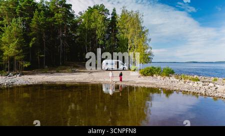 Un couple d'hommes et de femmes se tiennent sur la rive tranquille du lac Saimaa Finlande, entouré d'arbres luxuriants et d'une vue panoramique. Un camping-car est garé à proximité, invitant à l'aventure et à l'exploration. Banque D'Images