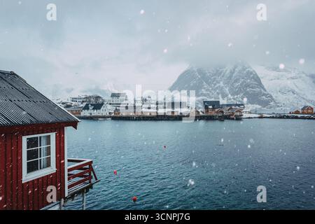 La neige tombe sur un village de pêcheurs norvégien, avec des cabanes rouges et des montagnes enneigées en arrière-plan Banque D'Images