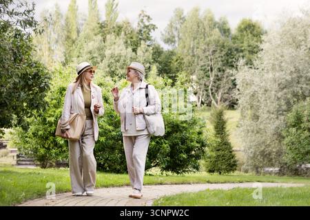 Portrait en longueur de deux femmes âgées marchant à l'extérieur dans des tenues élégantes beige clair et crème décontractées avec chapeaux, pantalons en lin, vestes, sac et Banque D'Images