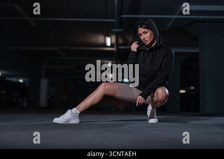 Dans un parking souterrain faiblement éclairé, une fille en sweat à capuche noir squats avec une jambe tendue. Elle respire la confiance et le style tout en mettant en valeur la mode de rue décontractée. Banque D'Images