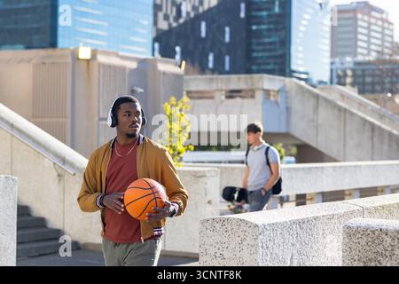Homme afro-américain portant une veste avec des écouteurs marchant sur la place avec basket-ball, espace de copie Banque D'Images