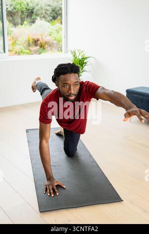 Homme afro-américain pratiquant le yoga d'équilibrage pose sur tapis de yoga noir dans le salon par fenêtre Banque D'Images