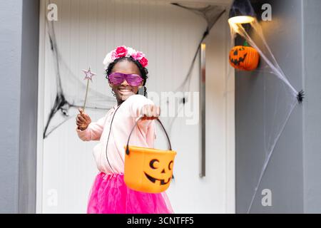 Fille afro-américaine portant tutu tenant seau de citrouille et baguette sur le porche avec des toiles d'araignée Banque D'Images