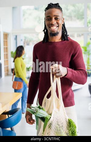 Couple diversifié portant des sacs d'épicerie en maille réutilisables remplis de légumes à feuilles dans la cuisine moderne Banque D'Images