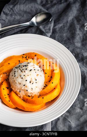 Riz collant à la mangue arrosé de sauce noix de coco dans un bol peu profond gros plan : dessert thaïlandais avec riz gluant, fruits frais et graines de sésame grillées Banque D'Images