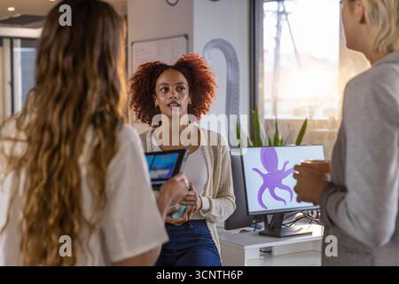 Diverses collègues féminines collaborant autour du bureau dans le bureau en utilisant la tasse à café de tablette, moniteur Banque D'Images