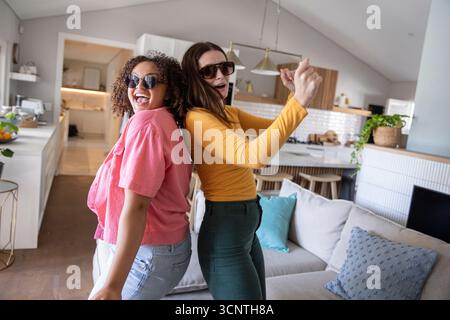 Diverses amies féminines portant des lunettes de soleil et dansant dos à dos près de l'îlot de cuisine et du canapé Banque D'Images