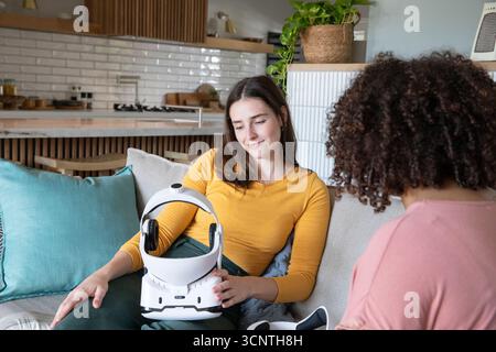 Divers amis féminins testant le dispositif de casque tout en étant assis sur un canapé avec des coussins près de la cuisine ouverte Banque D'Images