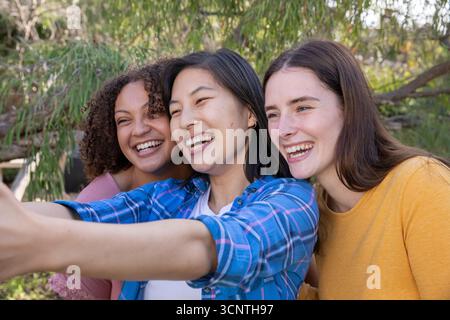 Diverses amies féminines prenant selfie avec smartphone tout en se tenant parmi les arbres ensoleillés au parc Banque D'Images