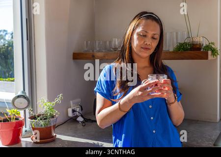 Femme asiatique debout au comptoir de la fenêtre, tenant une tasse de thé en verre, regardant la boisson Banque D'Images