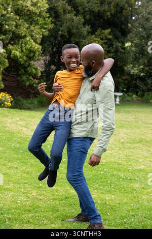 Père afro-américain et fils soulevant garçon sur la pelouse de l'arrière-cour près de fleurs jaunes, banc blanc Banque D'Images