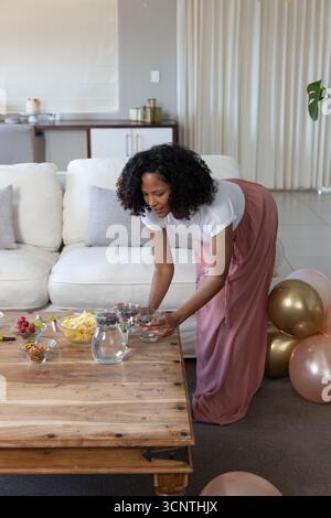 Femme afro-américaine préparant des collations et des boissons sur une table basse dans le salon, des ballons Banque D'Images