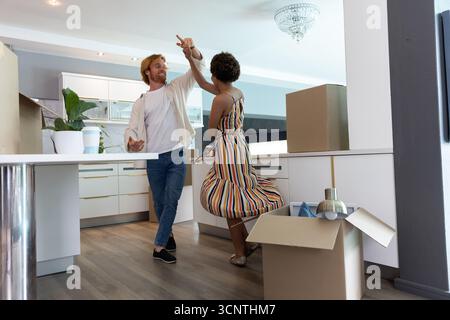 Couple diversifié dansant parmi les boîtes mobiles déballées dans la cuisine avec plante en pot, tasse à café Banque D'Images